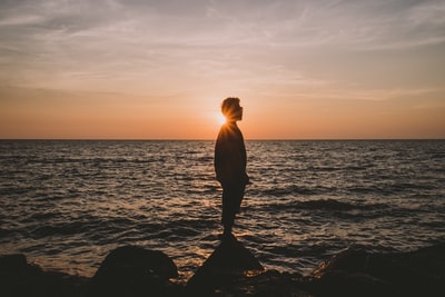 日落时，男人站在波浪形海滩旁的石头上的剪影